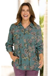 Langärmlige Bluse in grünblau mit Blumenmuster, Hemdkragen, Knopfleiste, locker geschnitten, getragen von lächelnder Frau, Hände in Taschen, kombiniert mit violetten Hosen