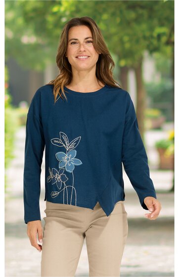 Marineblaues Langarmshirt mit großem hellblauem und weißem Blumendesign auf Vorderseite, Rundhalsausschnitt, asymmetrischer Saum mit Schlitz, getragen von lächelnder stehender Frau, legerer Schnitt