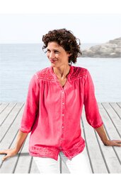 Rosa Bluse mit Spitzenbesatz an Schultern und Saum, durchgehende Knopfleiste, V-Ausschnitt, 3/4-ärmel, lockere Passform, getragen von sitzender Frau vor Meereskulisse