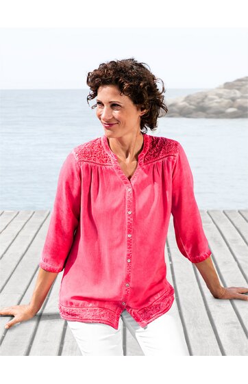 Rosa Bluse mit Spitzenbesatz an Schultern und Saum, durchgehende Knopfleiste, V-Ausschnitt, 3/4-ärmel, lockere Passform, getragen von sitzender Frau vor Meereskulisse