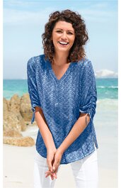 Blaues Damenoberteil mit floralem Muster, V-Ausschnitt, 3/4-Ärmel mit Krempelband, lockere Passform, getragen von lächelnder Frau am Strand stehend, weiße Hose