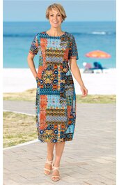 Midikleid mit buntem geometrischem Muster, dominant in Blau, Orange und Braun, rundem Halsausschnitt, kurzen Ärmeln, gerade geschnitten, getragen von stehender Frau am Strand, Seitentaschen