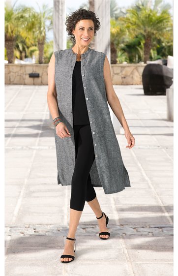 Graue lange Weste mit Druckknöpfen, ohne Ärmel, gerade geschnitten, leicht strukturiertes Material, getragen von stehender Frau mit schwarzem Oberteil und schwarzen Capri-Leggings, schwarzer Riemchensandale