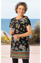 Schwarzes Kleid mit buntem Blumenmuster, kurze Ärmel, rundhals, gerade geschnitten, farbige Bordüre am Saum, getragen von stehender lachender Frau