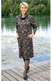 Knielanges Kleid mit Paisley-Muster in Schwarz und Weiß, langer Ärmel, hoher locker fallender Rundkragen, mit seitlichen Eingrifftaschen, getragen von stehender Frau auf Holzsteg vor See