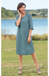 Kleid in graugrün mit feinem, leicht strukturiertem Muster, U-Boot-Ausschnitt, halblange Ärmel, knielang, lockere Passform, getragen von stehender Frau vor See und Wiese
