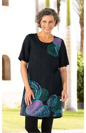 Schwarzes Locker sitzendes Kleid mit Rundhalsausschnitt, halblangen Ärmeln, abstraktem Kreismuster in Türkis und Lila, knieumspielende Länge, getragen von stehender lächelnder Frau