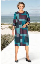 Kleid mit geometrischem Muster in Blau- und Türkistönen, Rundhalsausschnitt, 3/4-lange Ärmel, knielang, gerade geschnitten, getragen von stehender Frau vor Strandkulisse