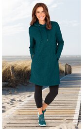 Dunkeltürkises Sweatkleid mit Kapuze, lange Ärmel, zwei seitliche Eingrifftaschen, sportiver Schnitt, getragen von stehender Frau am Strand, kombiniert mit schwarzer Capri-Leggings und türkisfarbenen Sportschuhen