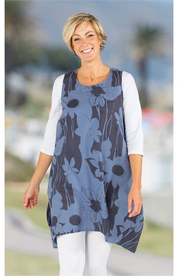 Ärmellose Tunika in dunklem Blau mit großem blauen Blumenmuster, Rundhalsausschnitt, asymmetrischer Saum, getragen von stehender lächelnder Frau mit weißen 3/4-Ärmeln und weißer Hose
