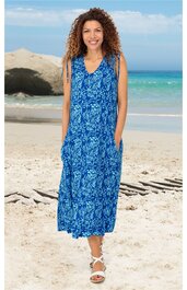 Blaues Maxikleid mit weißem Blumenmuster, V-Ausschnitt, ärmellos mit Bindebändern an den Schultern, seitliche Eingrifftaschen, gerade geschnittener Saum, getragen von stehender Frau am Strand, knöchellang