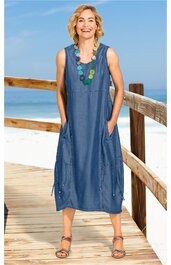 Ärmelloses midikurzes Kleid in Blau, unifarben, Rundhalsausschnitt mit dekorativem Besatz, seitliche Eingrifftaschen, seitliche Druckknöpfe, getragen von stehender Frau am Strand, lockere Passform