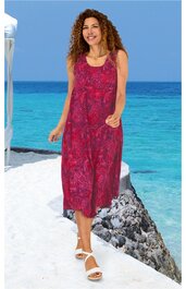 Ärmelloses midikleider in magenta mit rotem Blumenmuster, weiter Rundhalsausschnitt, seitliche Eingrifftaschen, ausgestellte Form, getragen von stehender Frau mit offenen Locken, weiße Sandalen, Strandkulisse