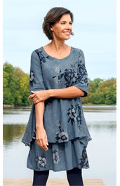 Blaugraues Kleid mit dunkelblauem Blumenmuster, rundem Halsausschnitt, dreiviertellangen Ärmeln, locker sitzend, mehrlagig, getragen von stehender Frau vor Wasser und Bäumen