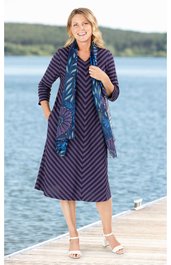 Knielanges Kleid in Dunkellila mit schwarzen diagonalen Streifen, Rundhalsausschnitt, 3/4-Ärmel, geschnitten in A-Linie, dazu blauer Schal mit floralem Muster, getragen von lächelnder stehender Frau auf Steg vor Wasser