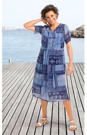 Blaues Patchwork-Midikleid mit verschiedenen Musterflächen, Kurzarm, V-Ausschnitt mit Knopfleiste, lockerer Schnitt, getragen von stehender Frau am Wasser auf Holzsteg, trägt weiße Sandalen