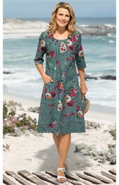 Kleid in grau-grün mit großflächigem Blumenmuster in Rot und Beige, Rundhalsausschnitt, Knopfleiste vorne, 3/4-Ärmel, seitliche Taschen, knielang, getragen von lächelnder Frau stehend am Strand