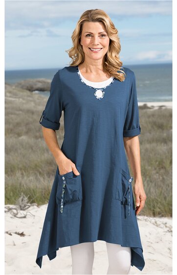 Blaues Longshirt mit weitem Schnitt, asymmetrischem Saum, Rundhalsausschnitt mit weißem Spitzeneinsatz, halbarmig, zwei aufgesetzte Taschen vorne mit Knöpfen, getragen von stehender Frau am Strand