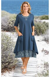 Blaues, knielanges Kleid mit floralem Muster am Saum, Rundhals, kurze Ärmel, zwei seitliche Taschen mit Kordeln, getragen von stehender Frau am Strand, weit geschnitten
