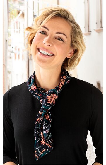Schwarzes Langarmshirt, rundhals, getragen von lächelnder Frau mit kurzen blonden Haaren, schwarzer Schal mit floralem Muster in Rosa und Blau locker um den Hals gebunden