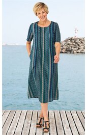 Knielanges Freizeitkleid in Dunkeltürkis mit senkrechten Streifen in Hellgrün, Blau und Beige, Rundhalsausschnitt, halblange Ärmel, gerade geschnitten, Taschen in der Seitennaht, getragen von gehender Frau auf Holzsteg vor Meer
