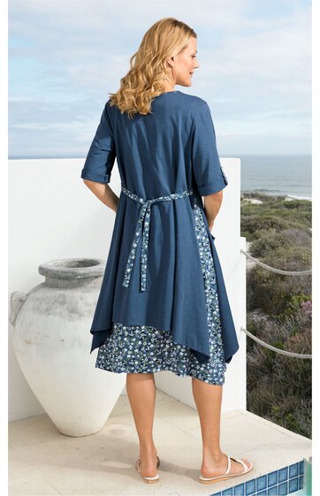 Dunkelblaues Freizeitkleid mit halbhohen Ärmeln, darunter hellblauer Rock mit weiß-blauem Blumenmuster, lockere Passform, Bindegürtel hinten, knieumspielend, getragen von stehender Frau mit Blick zur Seite