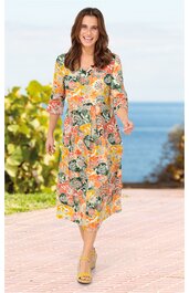 Midikleid in Beige mit buntem Blumenmuster, V-Ausschnitt, 3/4-Ärmel, tailliert, getragen von lächelnder Frau im Stehen, offene Sandalen, Hintergrund mit Meer und Pflanzen