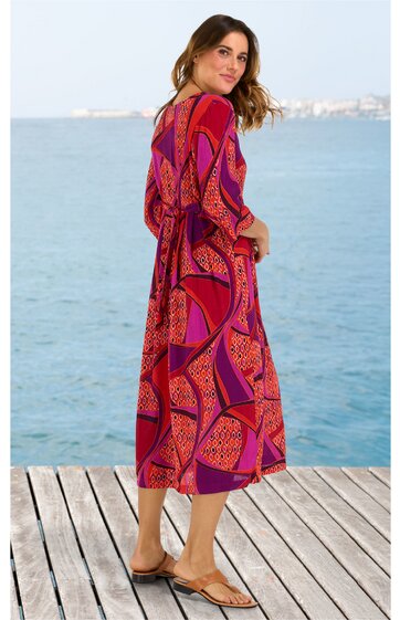 Midikleid in Rot und Dunkelviolett mit geometrischem Muster, V-Ausschnitt hinten, lange Ärmel, locker fallend, getragen von stehender Frau am Steg mit Blick zum Wasser, flache Sandalen