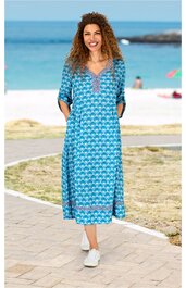 Blaues Maxikleid mit weißem geometrischem Muster, V-Ausschnitt mit Zierleiste, 3/4-Ärmel, gerade geschnitten, getragen von stehender Frau vor Strandkulisse