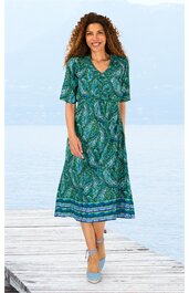 Midikleid in grün mit blauem Paisleymuster, V-Ausschnitt, halblange Ärmel, elastische Taille, breiter gemusterter Saum, getragen von stehender Frau mit lockigem Haar, Riemchenschuhe hellblau