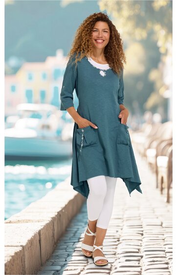 Türkisblaues asymmetrisches Longshirt mit 3‑4 Armeln, Rundhalsausschnitt mit weißem Einsatz, große aufgesetzte Taschen vorne, getragen von lächelnder stehender Frau mit weißen Leggings und weißen Sandalen