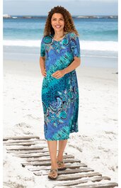 Midikleid in Blau- und Grüntönen mit abstraktem Muster, kurze Ärmel, Rundhalsausschnitt, locker sitzende Form, getragen von lächelnder Frau stehend am Strand, knielang