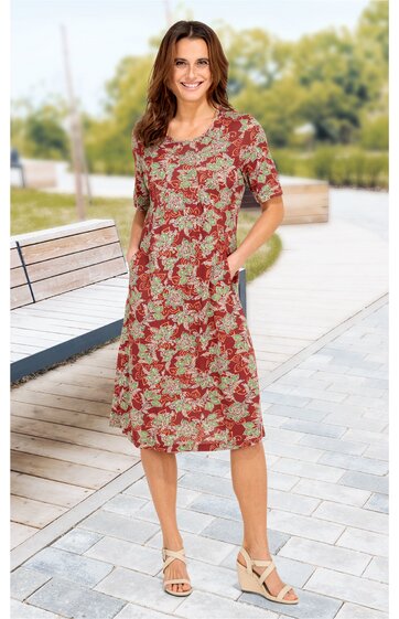 Kleid in rostrot mit beigem Blumenmuster, knieumspielend, halblange Ärmel, Rundhalsausschnitt, seitliche Taschen, getragen von stehender Frau auf Fußweg