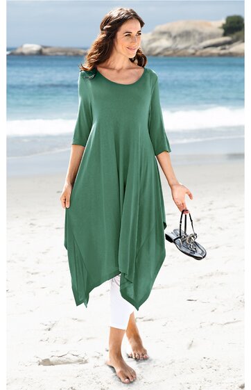 Locker geschnittenes asymmetrisches Kleid in grün, dreiviertellange Ärmel, rundhalsausschnitt, getragen von stehender Frau am Strand, mit weißer Leggings kombiniert, trägt schwarze Sandalen in der Hand