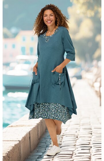 Blaugrünes Kleid, zweilagig mit ärmelloser, gemusteter Innenschicht und langer, unregelmäßiger, einfarbiger Außenschicht mit 3-4-Arm-Ärmeln, Rundhalsausschnitt, seitlichen Taschen, getragen von stehender Frau, knieumspielend