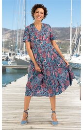 Midikleid in Blau mit rosa und schwarzen Blumenmustern, V-Ausschnitt, kurze Ärmel, lockere Passform, ausgestellter Rock, getragen von lächelnder Frau auf Steg vor Yachten, mit blauen Schuhen und Knöchelriemen