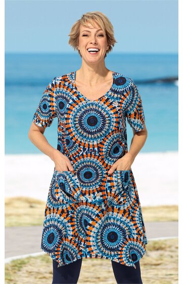 Kurzärmliges Kleid mit Rundhalsausschnitt, bunte kreisförmige Muster in Blau, Orange und Schwarz, zwei aufgesetzte Taschen vorne, knielang, getragen von lachender stehender Person am Strand