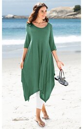 Locker geschnittenes asymmetrisches Kleid in grün, dreiviertellange Ärmel, rundhalsausschnitt, getragen von stehender Frau am Strand, mit weißer Leggings kombiniert, trägt schwarze Sandalen in der Hand