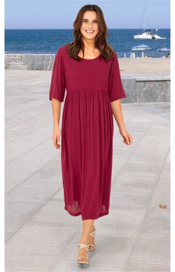 Bordeauxrotes Midikleid, einfarbig, rundhals, halblange Ärmel, tailliert mit gerafftem Rockteil, getragen von stehender Frau mit lockeren Haaren an Küstenpromenade