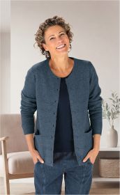 lächelnde Frau mit kurzen lockigen Haaren, trägt hellblaue Strickjacke mit Taschen und Knöpfen, dunkles Shirt und blaue Jeans, steht vor beigem Sessel und weißer Kommode mit Vase und Pflanze im hellen Raum