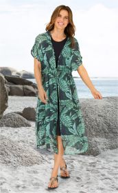 Frau am Strand trägt schwarzen Unterkleid mit darüber offenem, halblangem Viskose-Kaftan mit grünem, tropischem Blattmuster, schwarze Sandalen mit Schmuckringen, lächelnd, braune schulterlange Haare, heller Sand, Felsen und Meer im Hintergrund