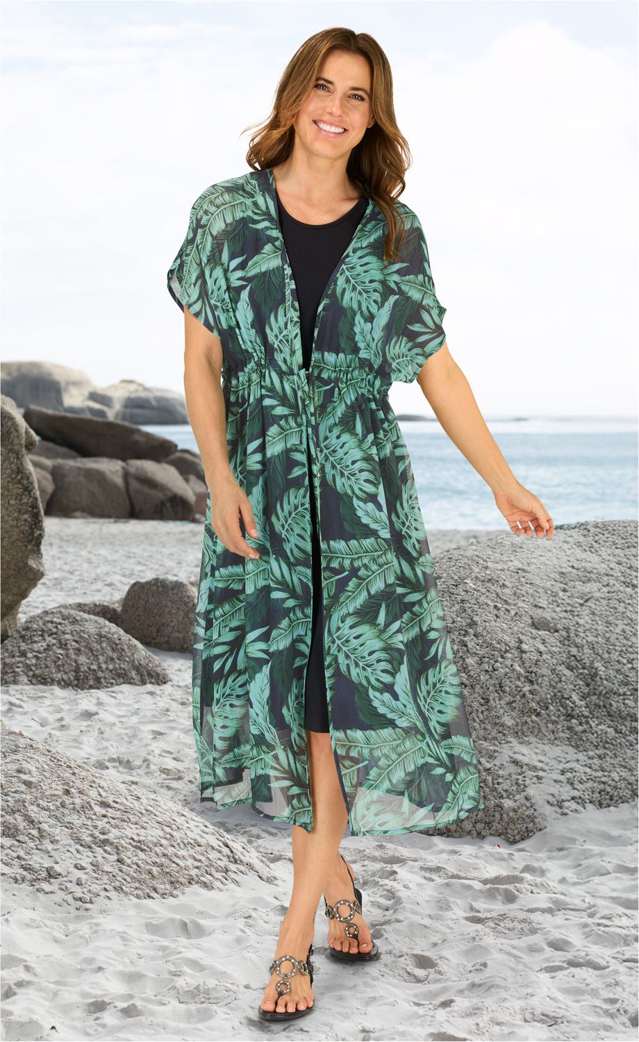 Frau am Strand trägt schwarzen Unterkleid mit darüber offenem, halblangem Viskose-Kaftan mit grünem, tropischem Blattmuster, schwarze Sandalen mit Schmuckringen, lächelnd, braune schulterlange Haare, heller Sand, Felsen und Meer im Hintergrund