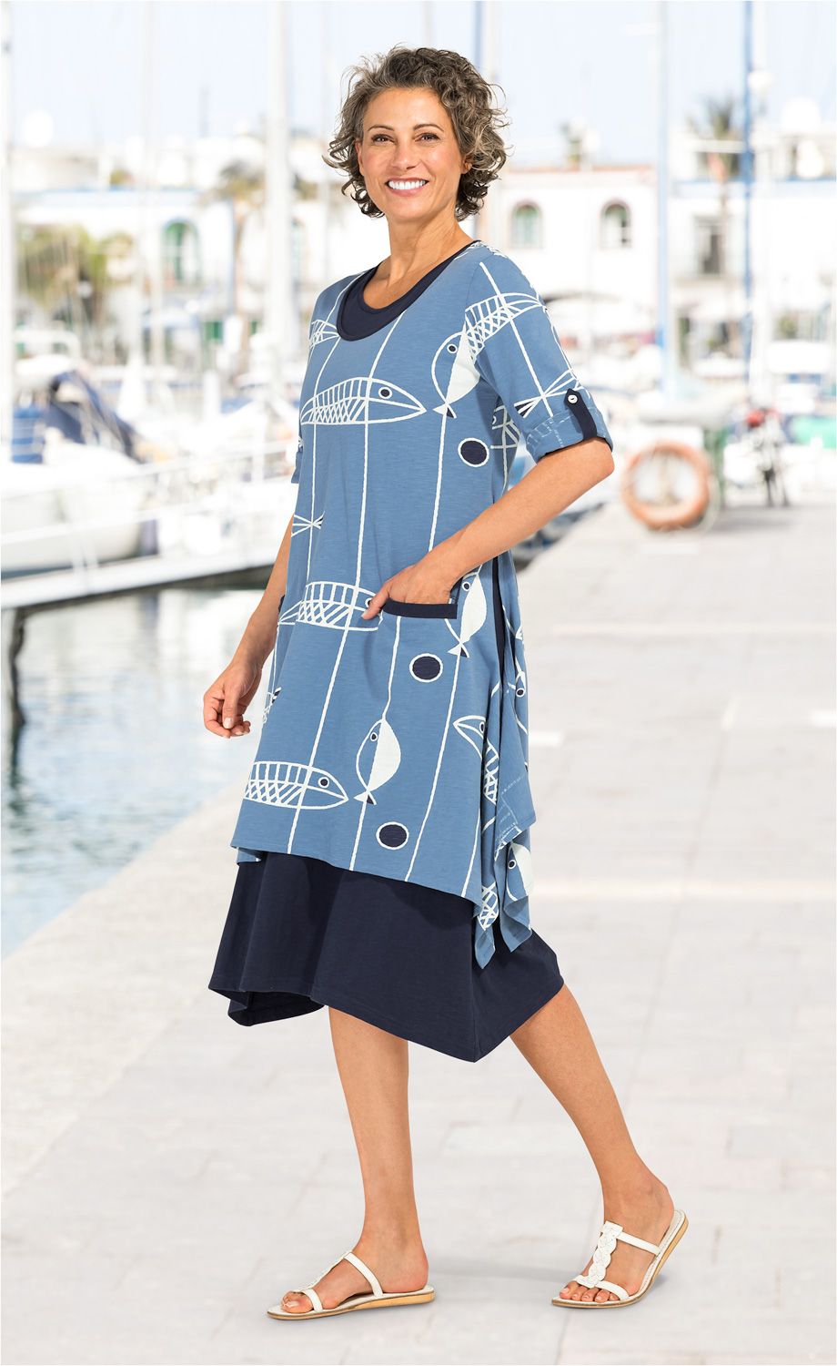 Frau trägt blaues Baumwoll-Kleid mit Fischmuster und drei Viertel Ärmeln, kombiniert mit dunkelblauem Unterkleid und weißen Sandalen, lächelnd am Hafen bei Sonnenschein