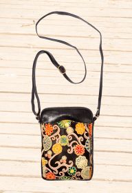 Kleine schwarze Leder-Tasche mit verstellbarem Schulterriemen, frontaler Außentasche mit farbenfrohem floralen Muster in Rot, Gelb, Grün und Beige, goldfarbener Druckknopfverschluss, strukturierter Hintergrund aus hellem Holz