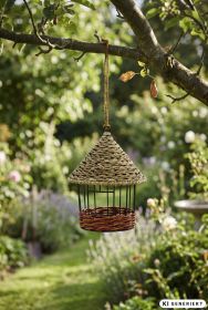 Vogelhaus aus geflochtenem Naturmaterial mit konischem Dach, hängend an Baumast im sonnigen Garten mit grünen Pflanzen und unscharfem Hintergrund aus Rasen, Blumen und Sträuchern.