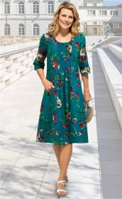 Frau trägt knielanges Baumwoll-Mix-Kleid mit floralem Muster in Blau, Grün und Rot, 3/4-Ärmel mit Umschlag, geht auf hellem Pflasterweg, hält Strohhut, trägt weiße Sandalen, lächelt, hellblonde Haare, sonniger Tag