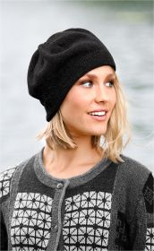Frau mit kurzen blonden Haaren trägt schwarze Alpaka-Mütze und graue Strickjacke mit weiß-schwarzem Muster, schaut lächelnd seitlich, unscharfer Wasserhintergrund