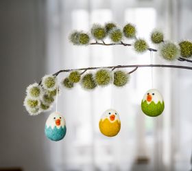 Zweig mit grünen, flauschigen Blüten, daran drei farbige Filz-Hänger Küken in den Farben Türkis, Gelb und Grün, weiße Köpfe mit orangenem Schnabel und schwarzen Augen, an dünner Schnur hängend
