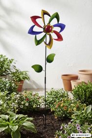 bunter Deko-Gartenstecker in Blumenform mit rot, gelb, grün und blau lackierten beweglichen Blütenblättern, grüne Blätter am schwarzen Metallstab, im Beet mit verschiedenen Pflanzen und Blumen, Terrakotta-Töpfe im Hintergrund