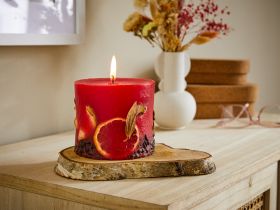 Brennende rote Stumpenkerze mit getrockneten Orangenscheiben, Kaffeebohnen und Blättern verziert, steht auf rustikalem Holzbrett auf hellem Holztisch vor Vase mit Trockenblumen, braunen Boxen und Brille.